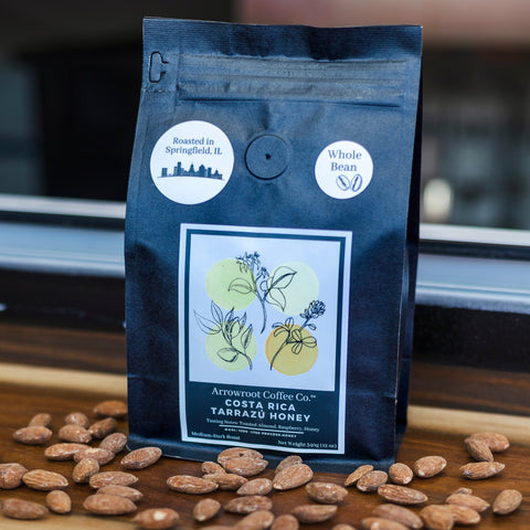 A bag of Costa Rica Tarazzú honey process coffee with roasted almonds beans scattered in front, set against a wooden background.